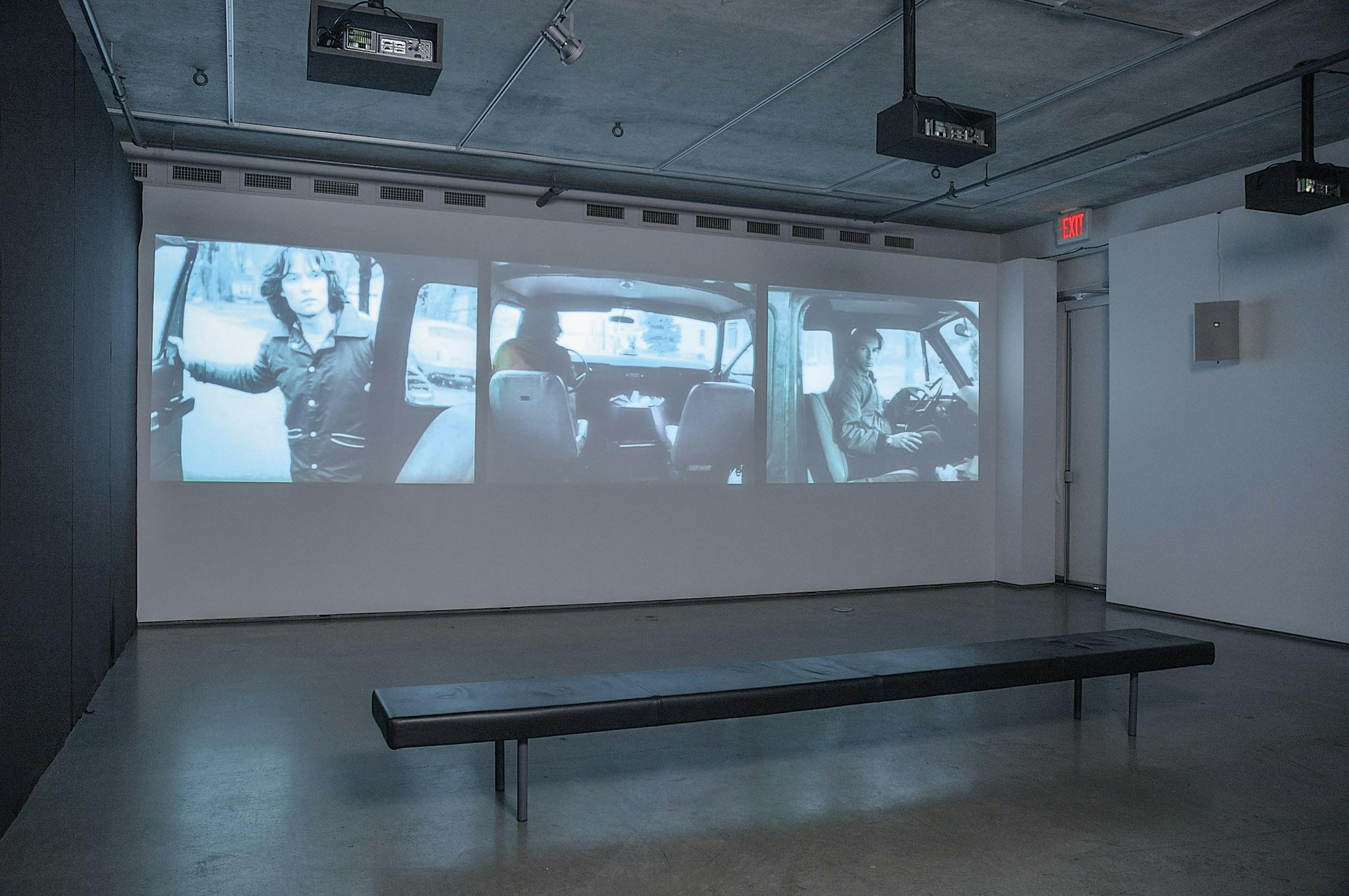 An installation image of three-channel video work projected on a gallery wall. The videos show people in a car from different angles. The left video show an image of a person standing outside while keeping one of the front doors of the car open.