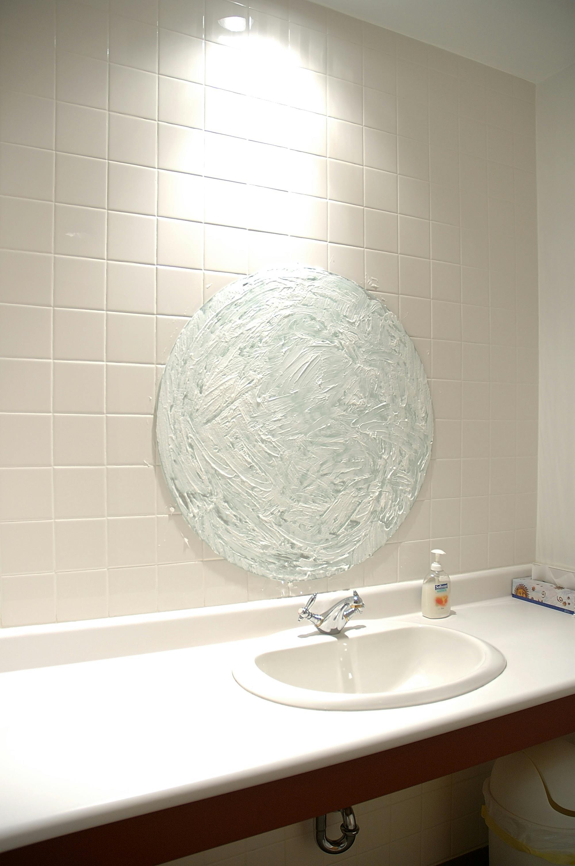 An installation image of a circular-shaped mirror above a sink. The mirror is entirely covered with a white soap-like form. 