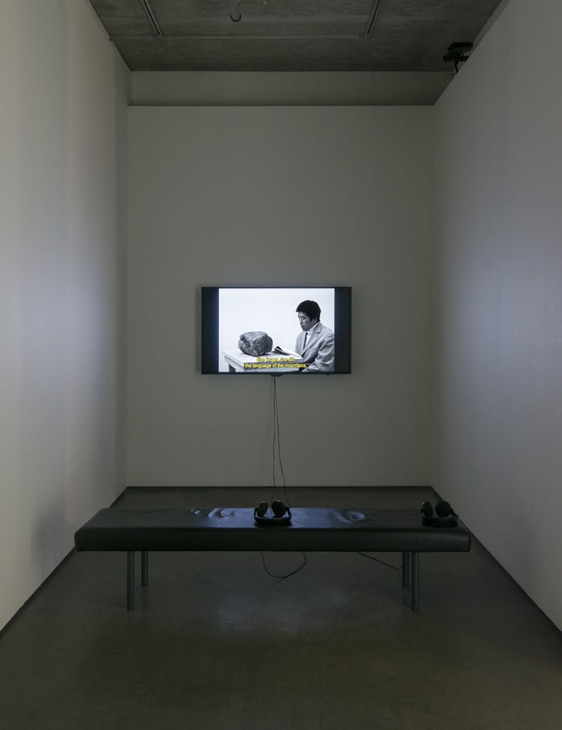 A monitor on a gallery wall displays a black and white video piece. The video depicts a person in a suit, reading a book, with a rock on a table in front. 