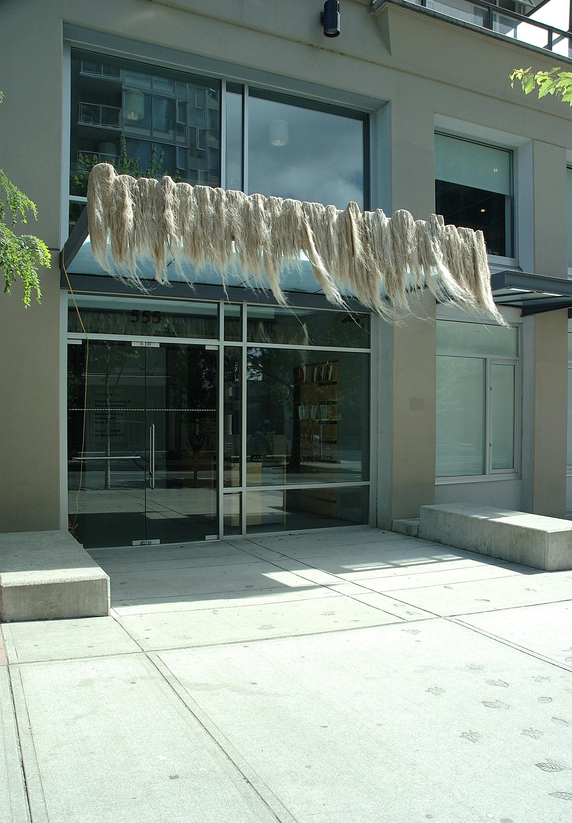 An installation image of an art piece above the CAG’s entrance door. On the glass-and-metal awning above the entrance, thick, ragged plumber's hemp are mounted. Strings of hemp lined up and swinging in the air may remind a viewer of long blond hair.