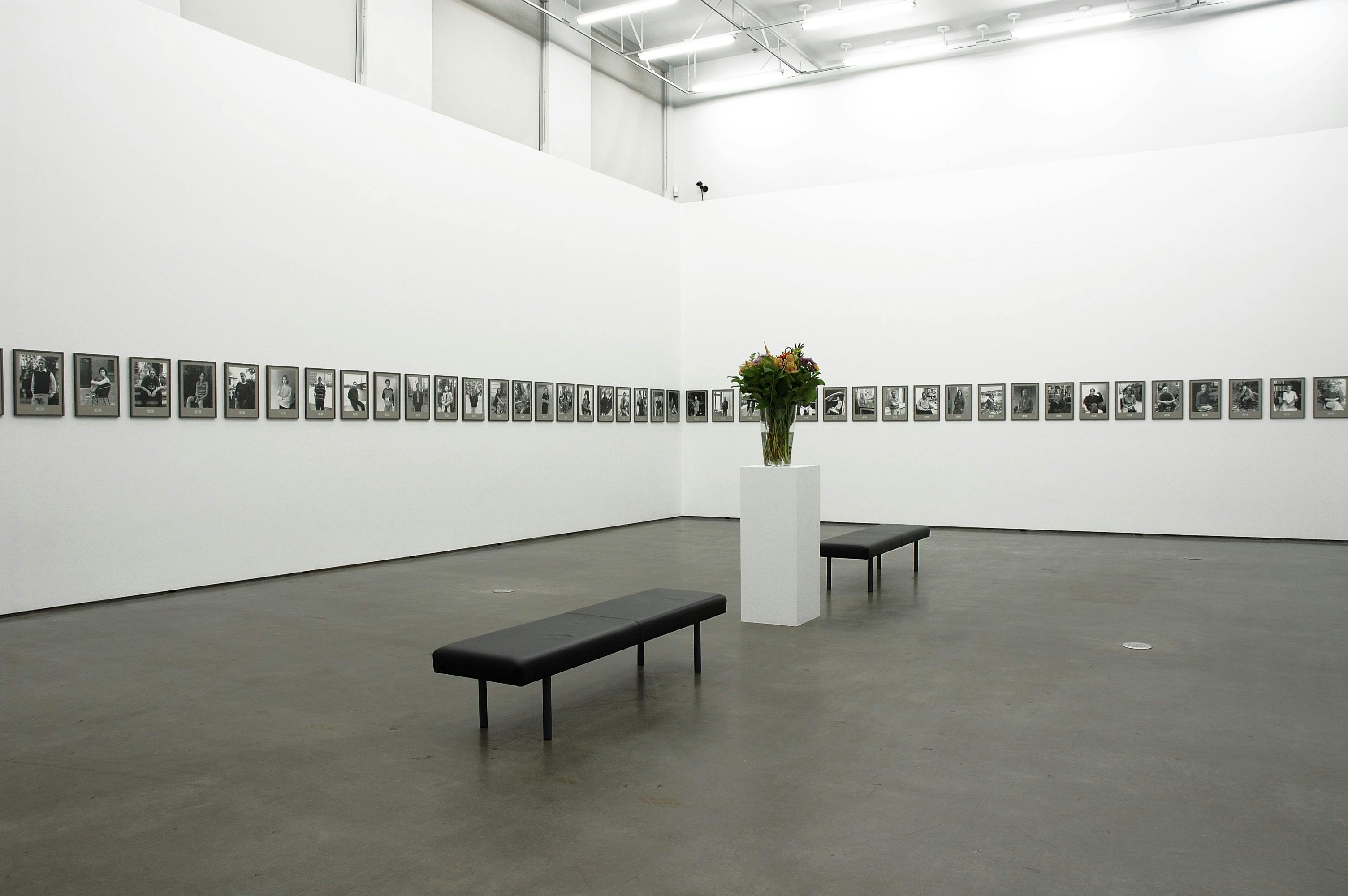 In a gallery, there are dozens of framed black and grey photos on the walls. At the centre of the space, there is a white plinth with pink flowers, with small black benches on either side of it.