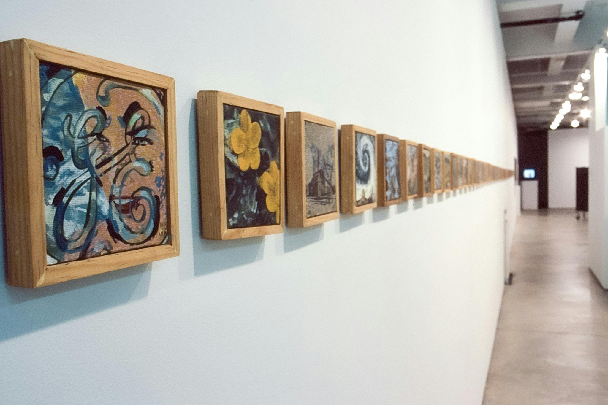 Multiple small-sized wooden framed paintings are mounted on the gallery wall. The one in the front depicts calligraphic lines that form a shape that resembles the capital letter H in a circle.