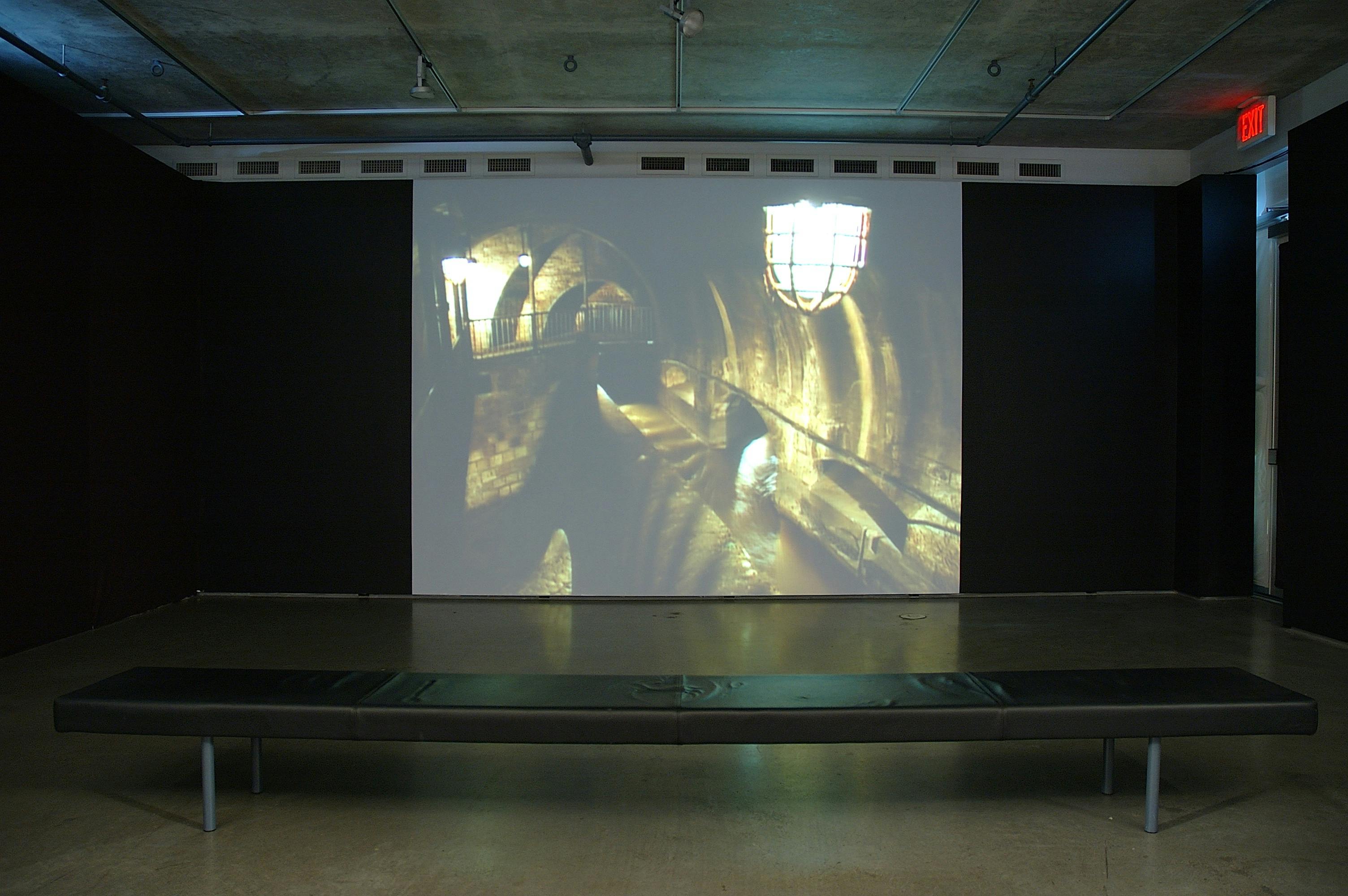An installation image of a video projected on a wall in a dimly lighted gallery. The video shows an aerial view of a sewer.