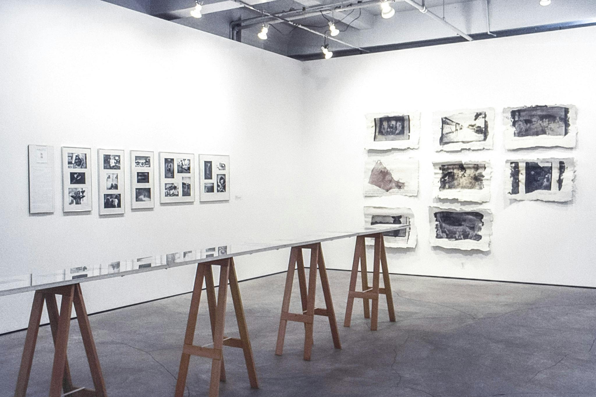 A gallery with several framed works mounted on the walls. In the centre of the room, there is a table-like form with a narrow, glossy top.  It is resting on 5 sets of A-shaped wooden table legs.