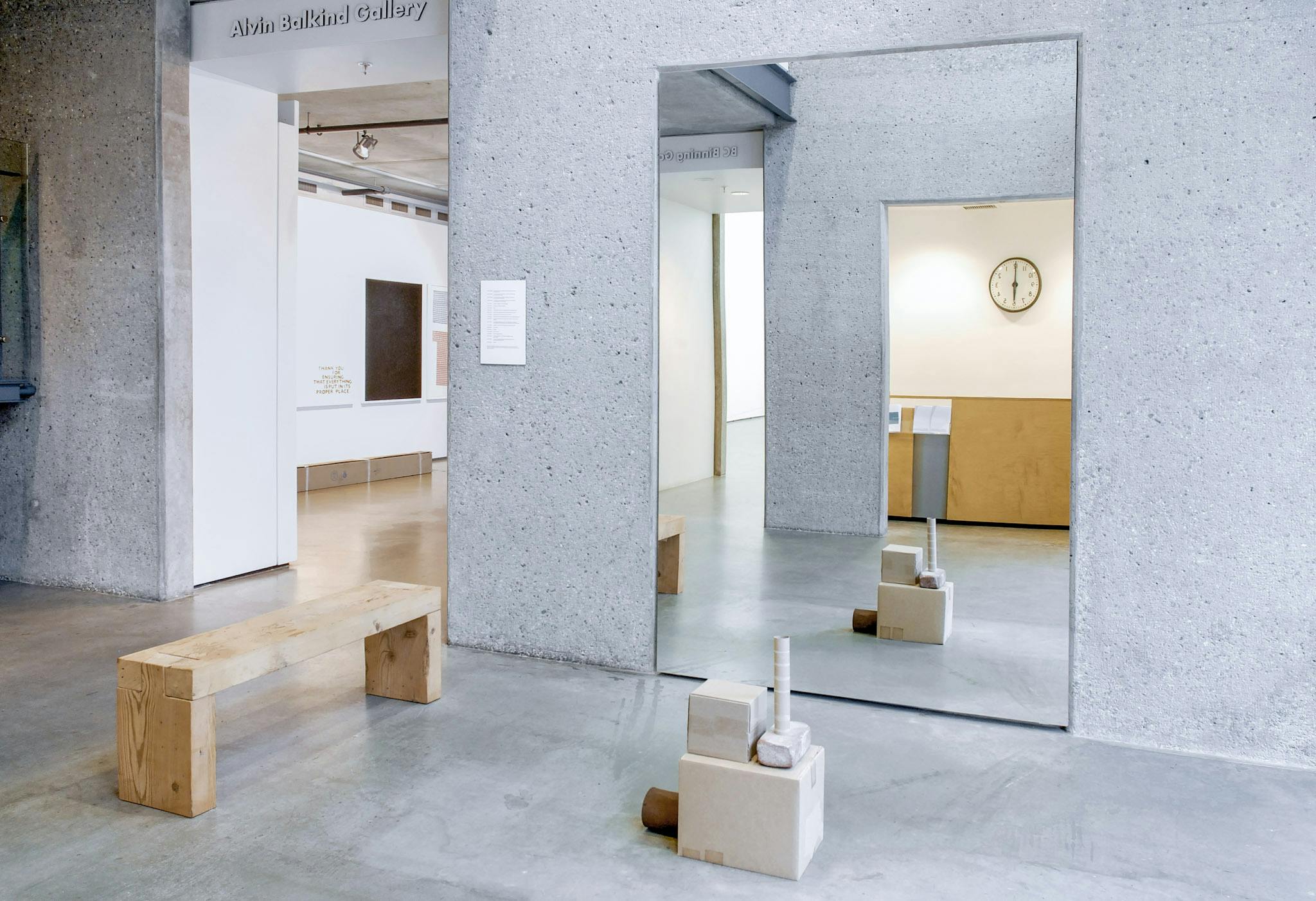 A large mirror is installed on the grey wall outside the Belkind Gallery. A wooden sculpture that looks like a stack of children’s toys is placed in front of that mirror. 