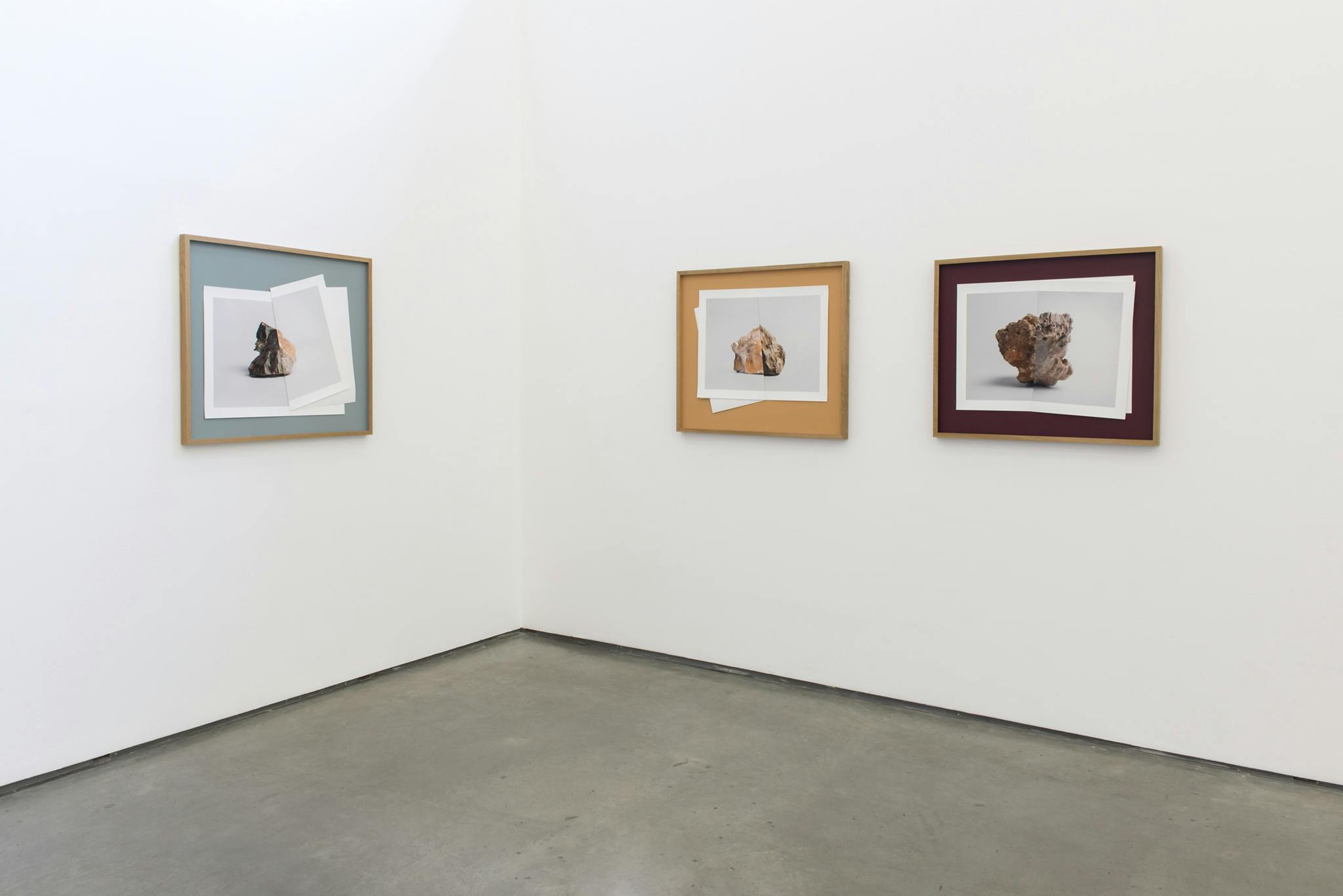 Three framed photographs of rocks hang in a gallery space. Each frame contains two folded photographs of rocks that connect at the fold to create an image of one, composite rock.