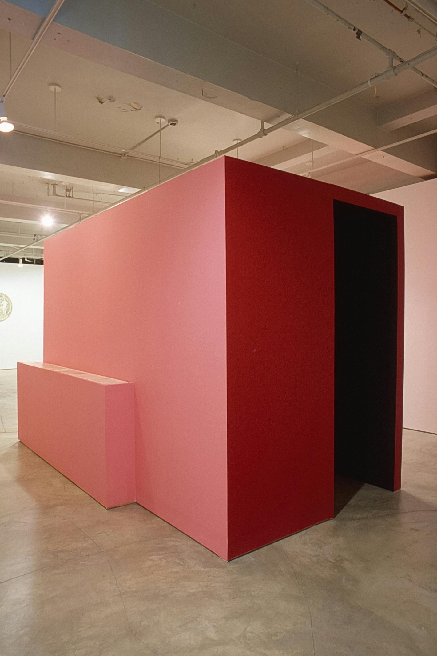 A large-sized red cube is built in a gallery space. A black rectangular on the right end of the cube looks like a door through which the visitors may walk inside the cube.