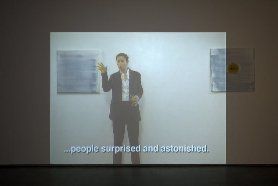 A projection on a gallery wall overlaying two paintings. The projection shows a person with their hand partially raised, giving a talk. The subtitles reads “people surprised and astonished.” 