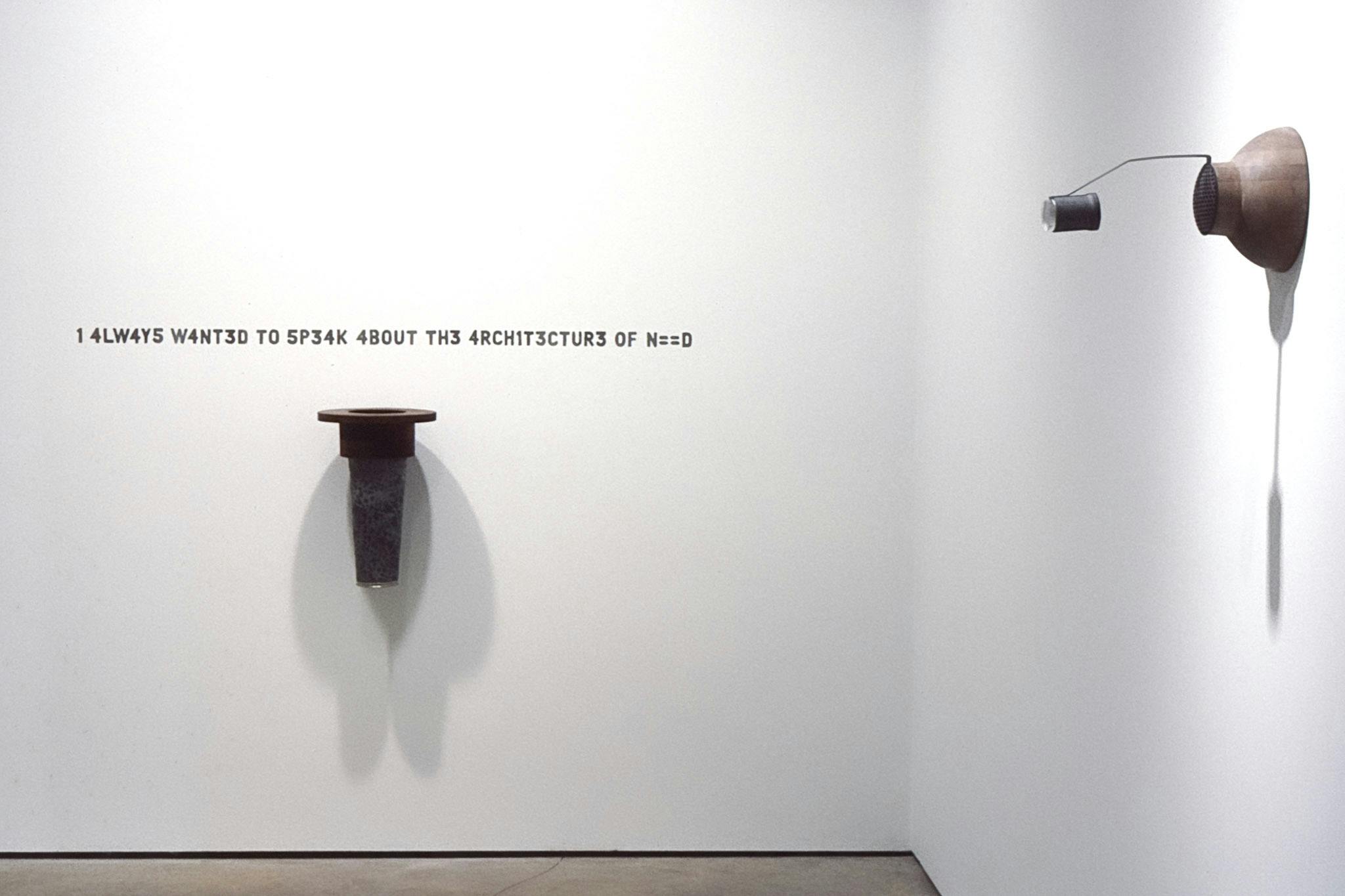 Two artworks are mounted on the gallery walls. Both are wooden sculptures, and one on the left has a sentence, “1 4LW4YS W4NT3D TO SP34K 4BOUT TH3 4RCHIT3CTUR3 OF N==D” typed on the space above it.