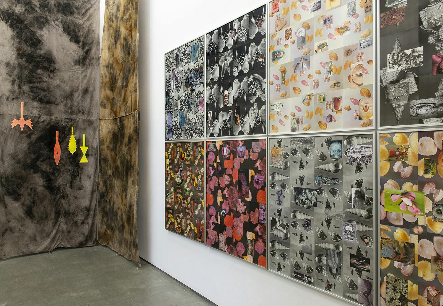 Eight framed prints of collages hang on a wall on the right side of the image. On the left there is brown tie dyed fabric hanging from the ceiling. Suspended from the fabric are tool-like sculptures.