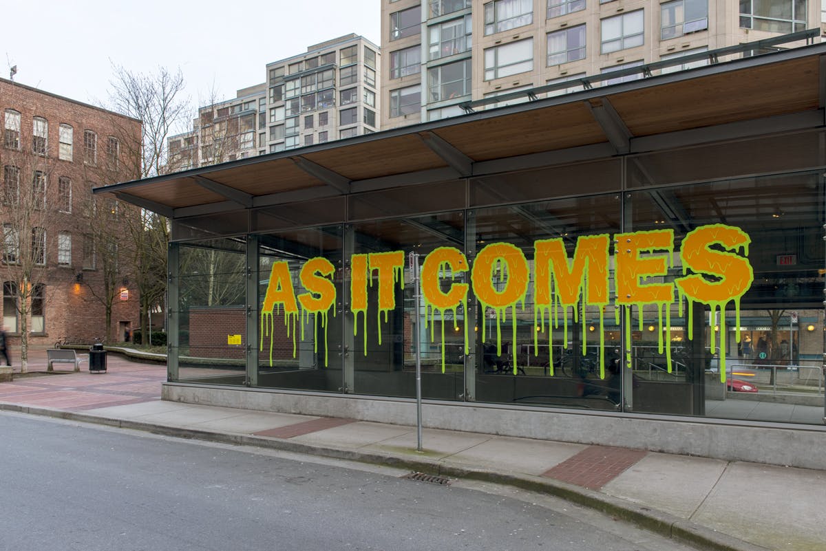 Text in vinyl installed on the glass exterior of Yaletown-Roundhouse Station. “AS IT COMES” appears in orange-yellow colour. Letters are outlined by dripping, neon green lines.