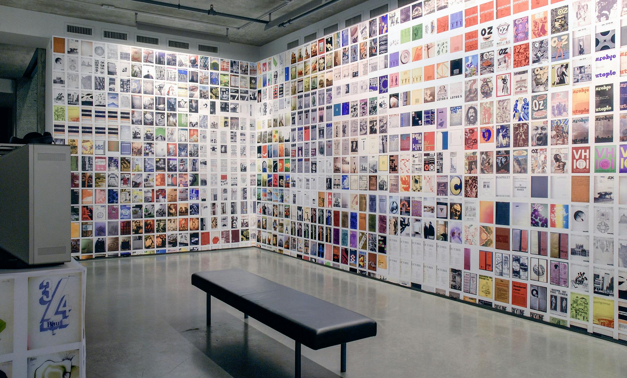 Printed images of different magazines entirely cover two walls of the gallery space. Most of those images are coloured. A black bench is placed in front of these two walls. 