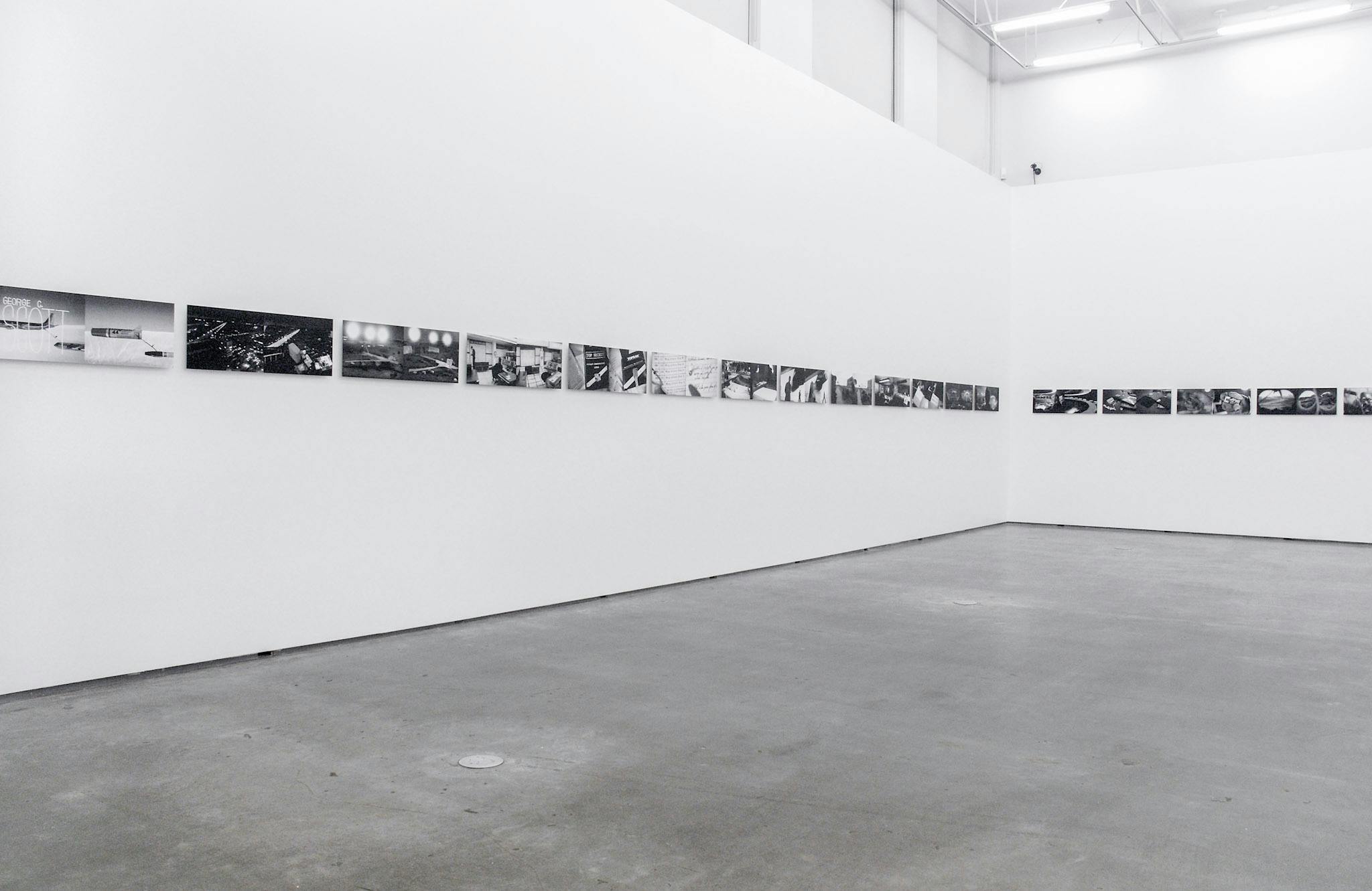 A number of same-sized black and white photographs are installed on the gallery walls. Two similar images are paired and exhibited side by side. Many of those images depict landscapes. 