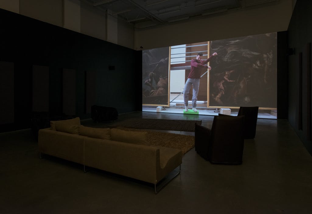 A large screen on a gallery wall in front of a sofa, rugs and chairs. A single-channel video is projected on the screen, depicting a person standing on a green block between two large oil paintings.