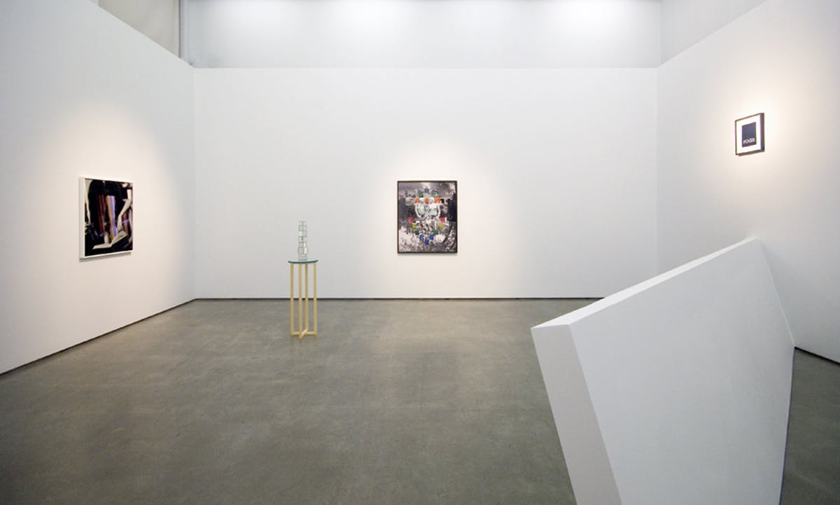 Installation shot of Elizabeth Zvonars work in a gallery. Three images hang on a different wall. A small table with cubes on top sits to the left, a large and long rectangular block sits to the right.