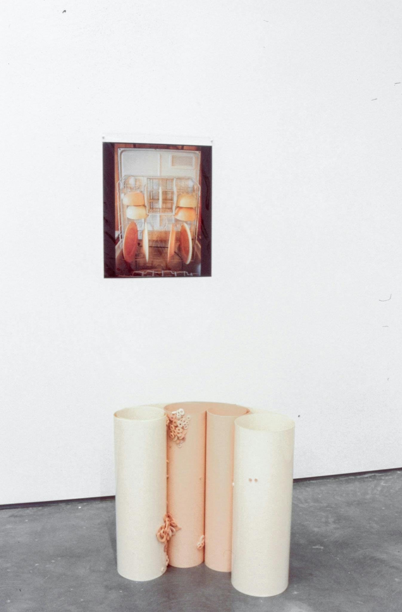 A photograph depicting an open dishwashing machine is installed on a gallery wall. Below this photograph, a sculpture made of four cylindrical-shaped objects sits on the floor. 