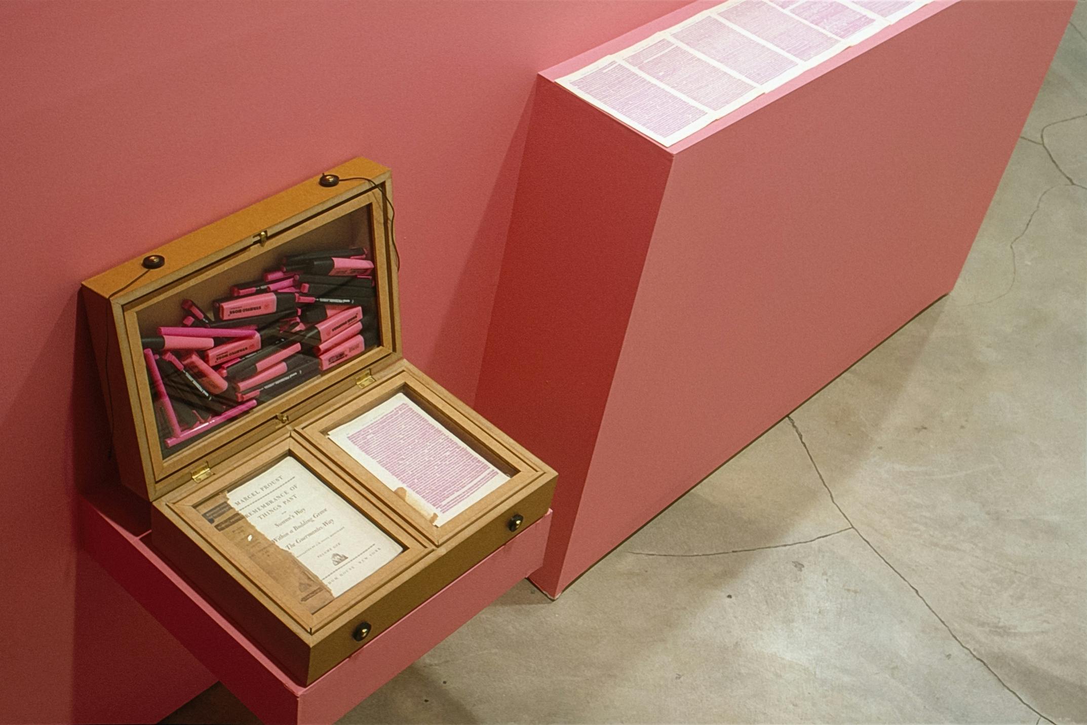 A small wooden cabinet is installed on a pink gallery wall. One of the containers is filled with pink markers used by the artist to highlight the texts in Marcel Proust's Remembrance of Things Past.