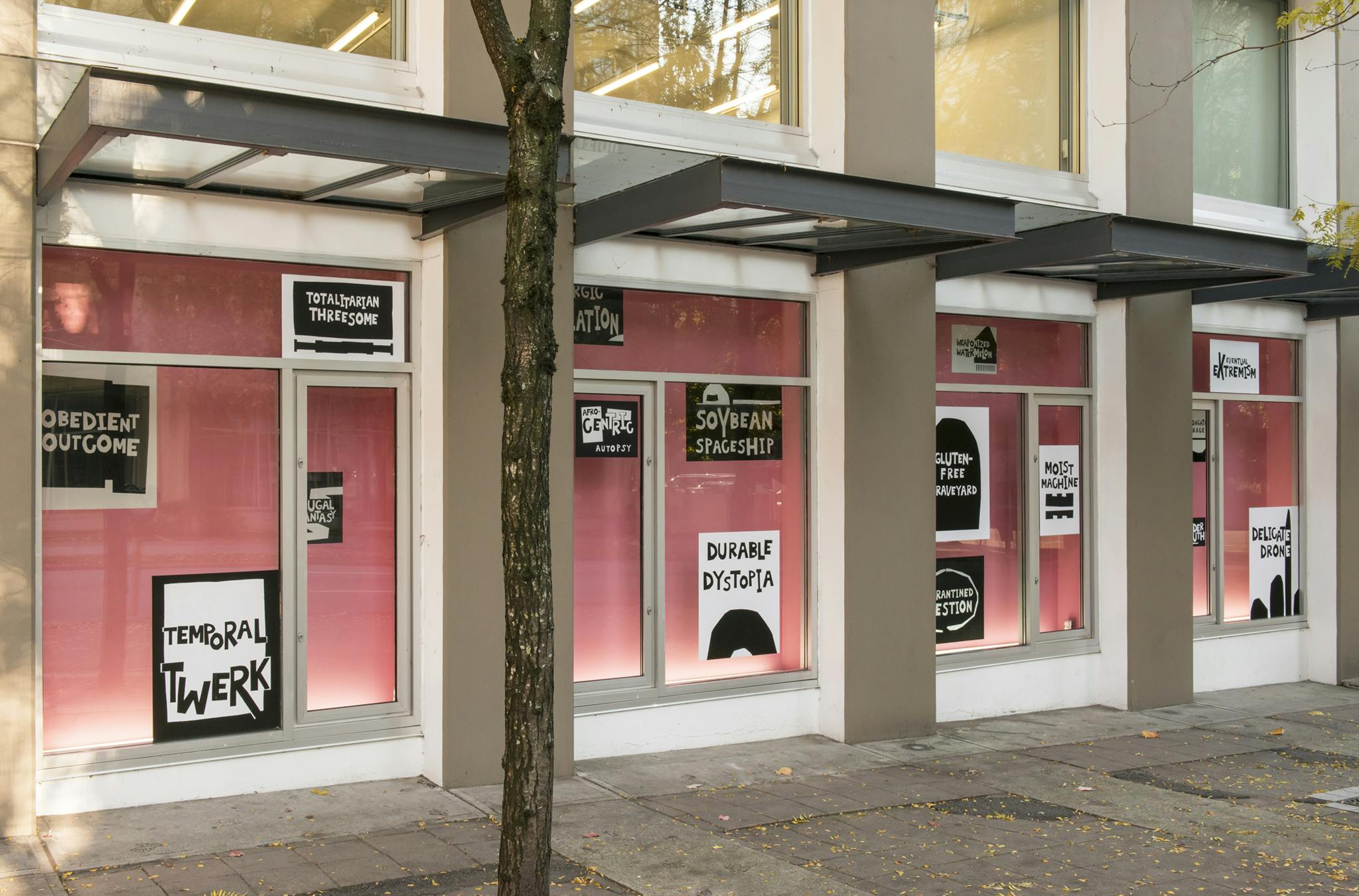 Four ground level façade windows at CAG installed with the work of Kameelah Janan Rasheed. Each window has a pink backdrop with black and white text-based vinyl prints scattered across.