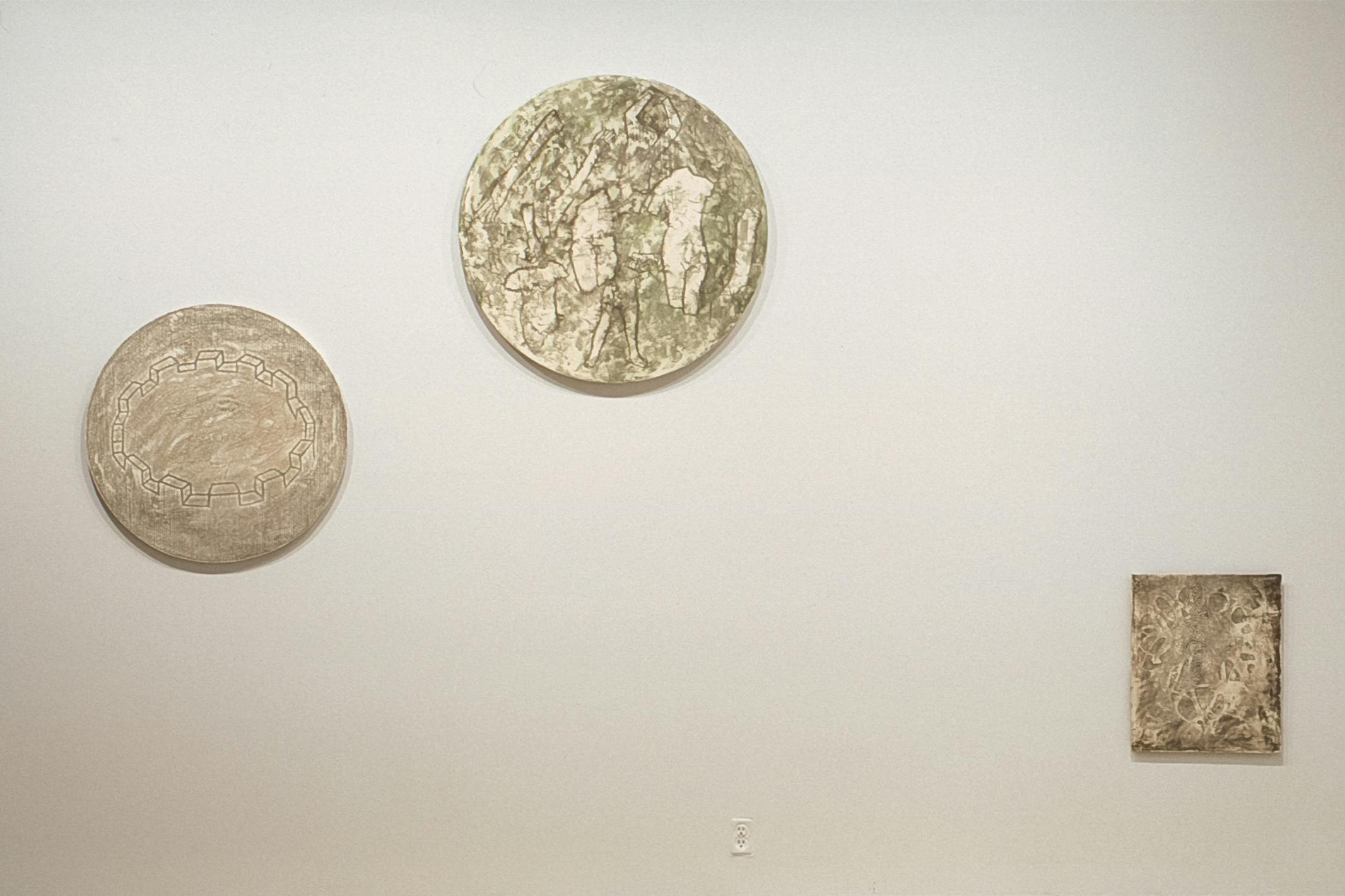 Three figures are installed on the gallery wall. All of them have dusted white surfaces, on which some drawings are made. Two pieces at the top are circular and the bottom one is rectangular. 