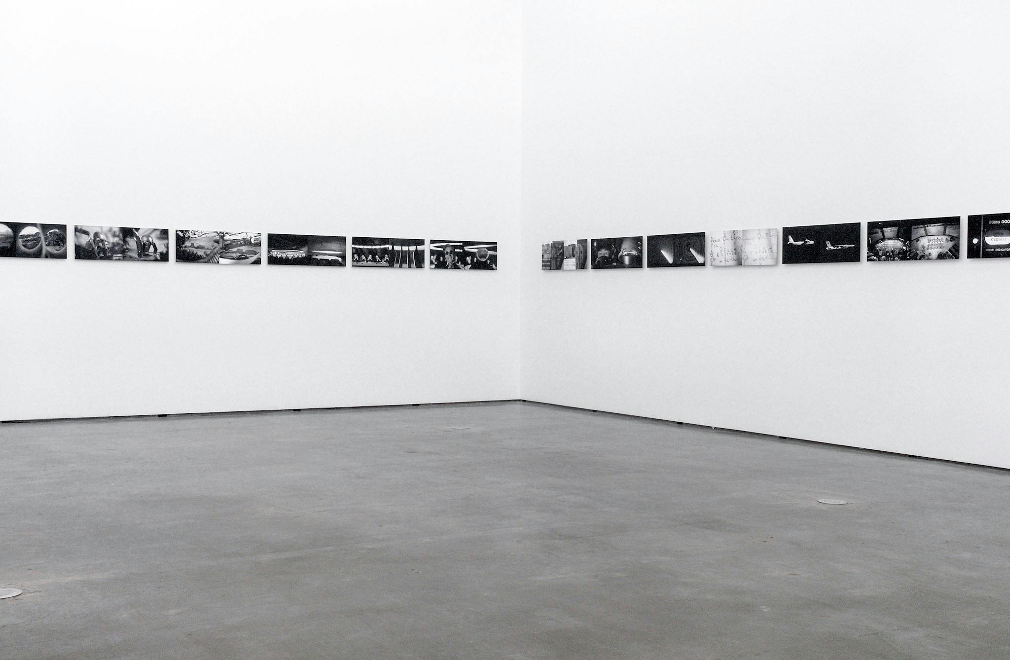 A number of same-sized black and white photographs are installed on the gallery walls. Two similar images are paired and exhibited side by side. Some of those images depict cityscapes.