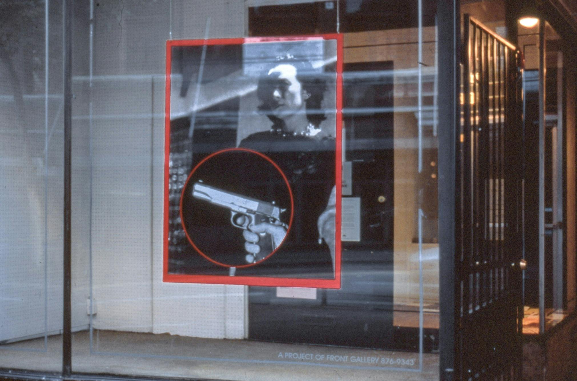 A large photo print hangs in a glass display on a street front. The photo has a red border and shows a person with their hand on their hip. In front of them, a closeup of a handgun in a red circle. 