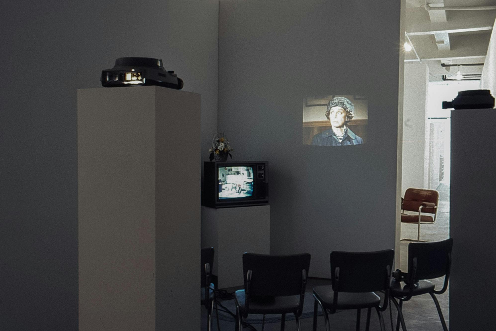 In the dark corner of a gallery space, there are two projectos and a small TV on plinths. Each of the devices plays a different video. Four chairs sit between the plinths, facing the projections.