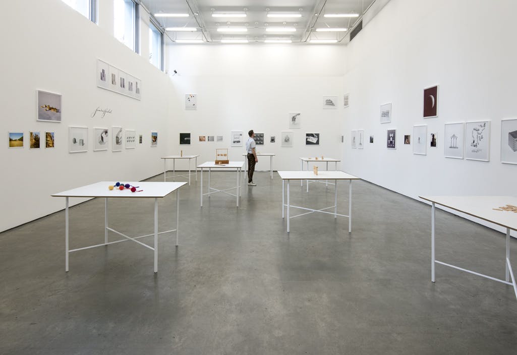 Installed in a gallery are various framed photographs and drawings and seven objects on white tables. A person stands amidst the tables looking up at one wall of framed works. 