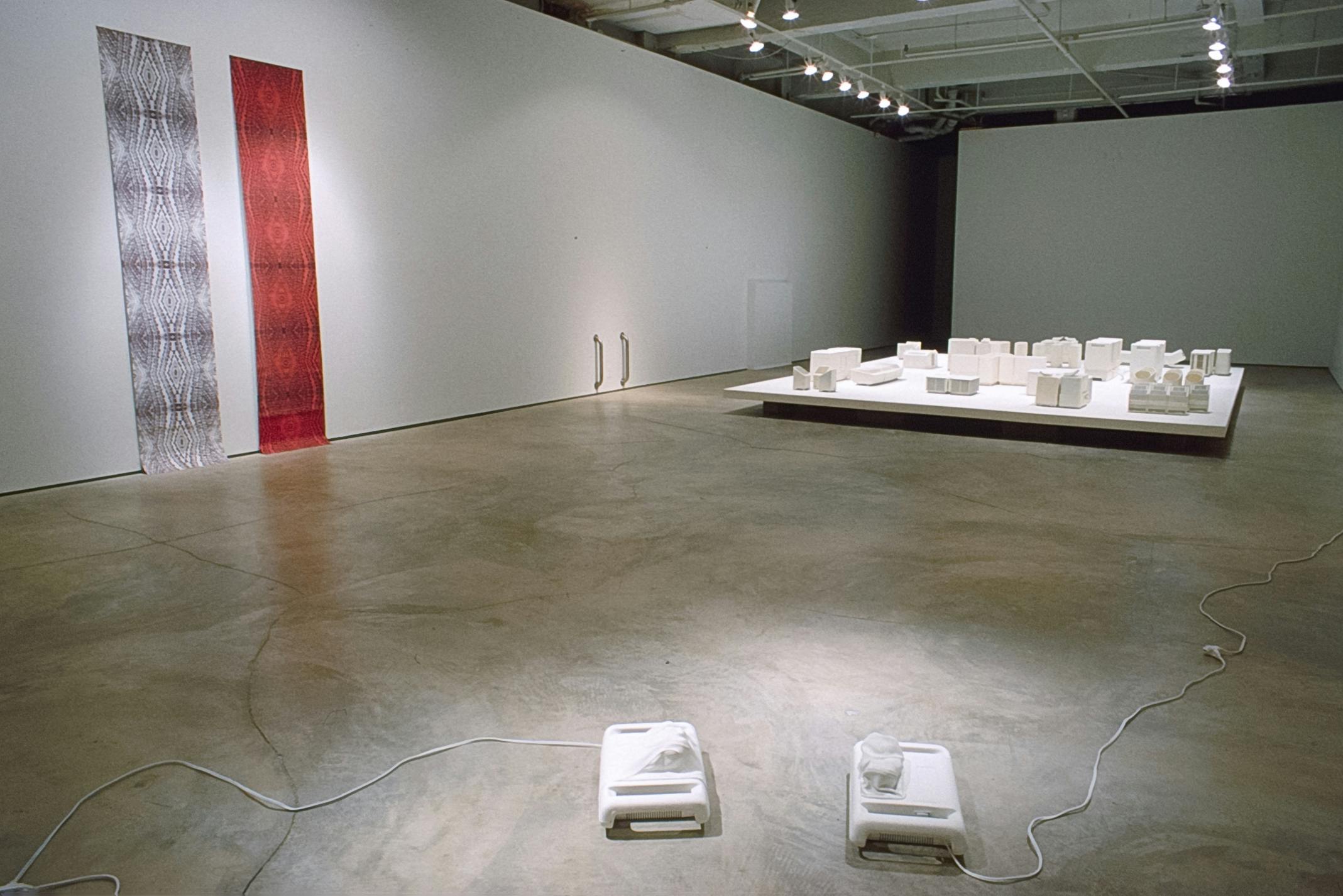 Several artworks are installed in the gallery. White miniatures of electronics are installed in the back. Two small white machines are connected to the cable and placed on the floor. 