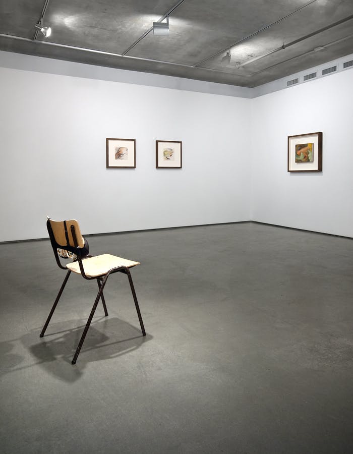 Three framed illustrations hang on two gallery walls. A chair sits on its own in the middle of the room with a bra attached to the back support. The bra is holding two round objects. 