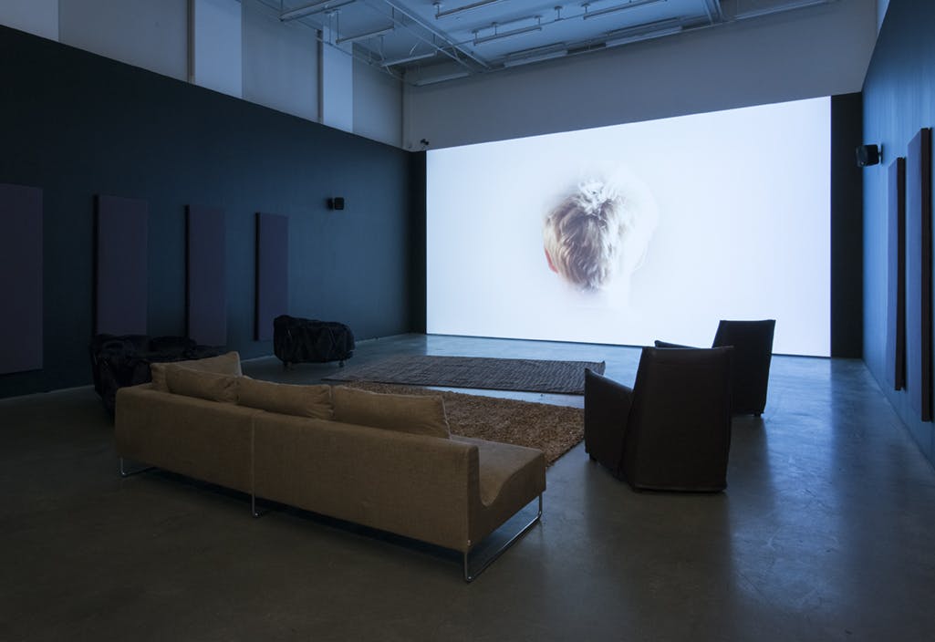 A large screen on a gallery wall in front of a sofa, rugs and chairs. A single-channel video is projected on the screen, depicting the back of a child’s head, with short blonde hair. 