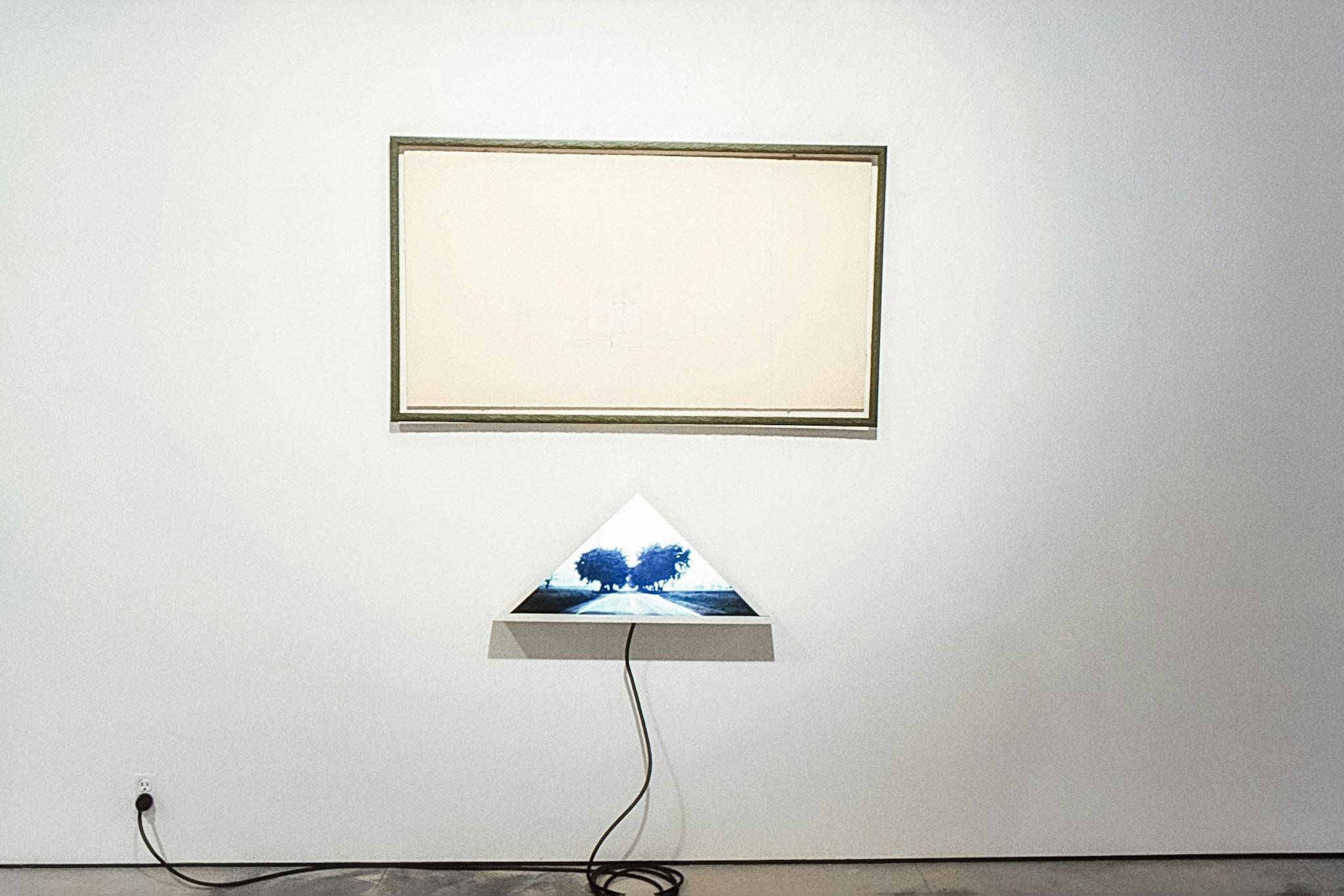 Two objects mounted on the wall of a gallery. One is a pale-yellow paper in a dark-green rectangle. Below, there is a light box in the shape of a triangle, showing an image of a wide road with trees.