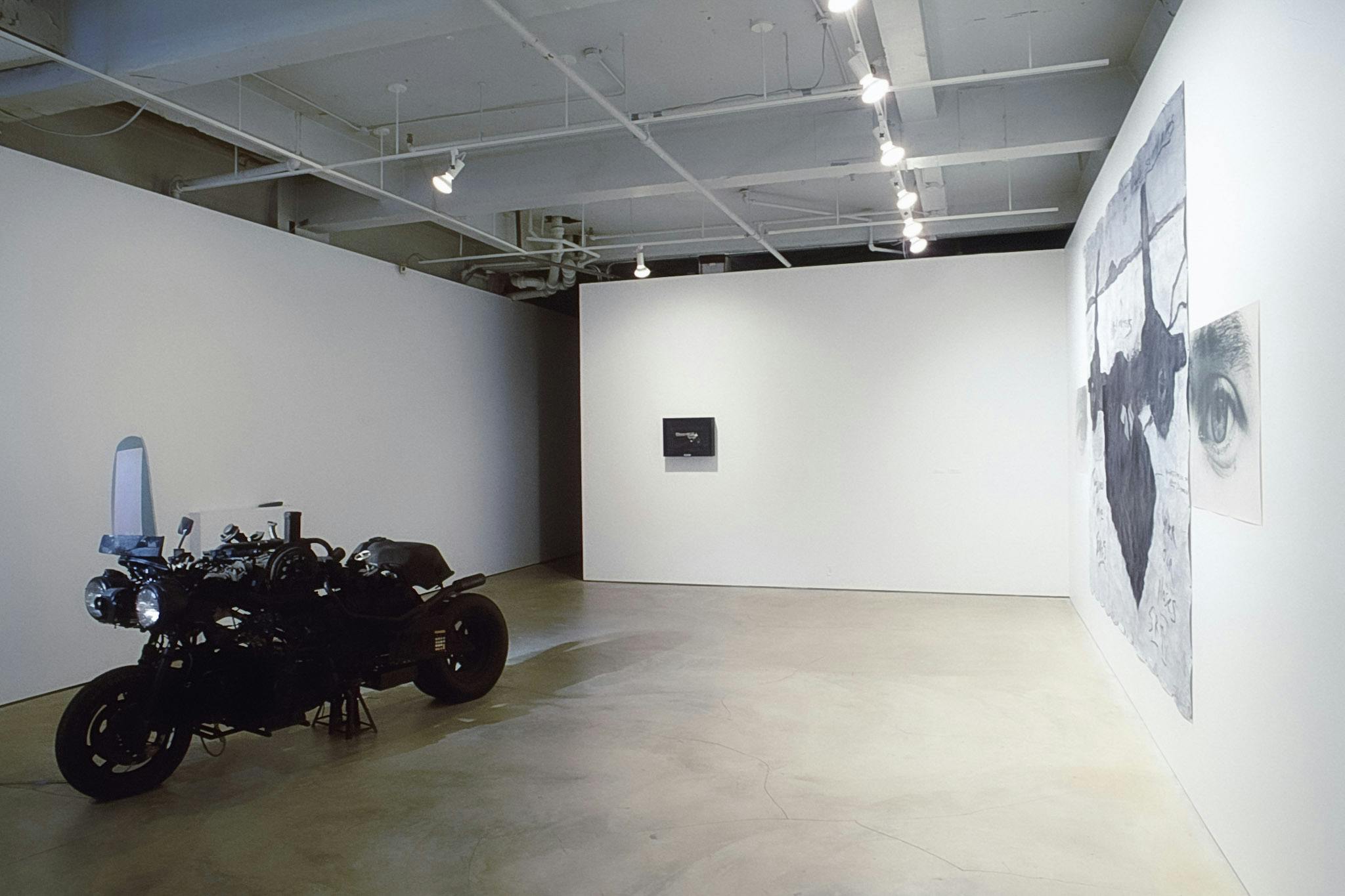 A black motorbike, a drawing of black airplane in between a pair of black and white photographs of human eyes, and a silver gun are installed in a gallery space. 