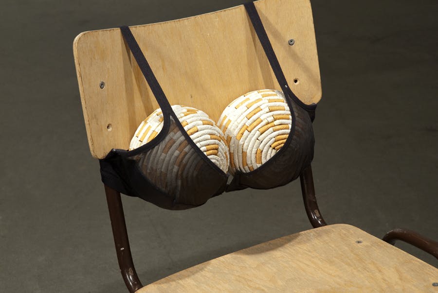 Detail shot displaying a chair with a black bra clasped on the back support of the chair. Two balls are held inside the bra. Both balls are covered by white and orange rolled paper resembling cigarettes.