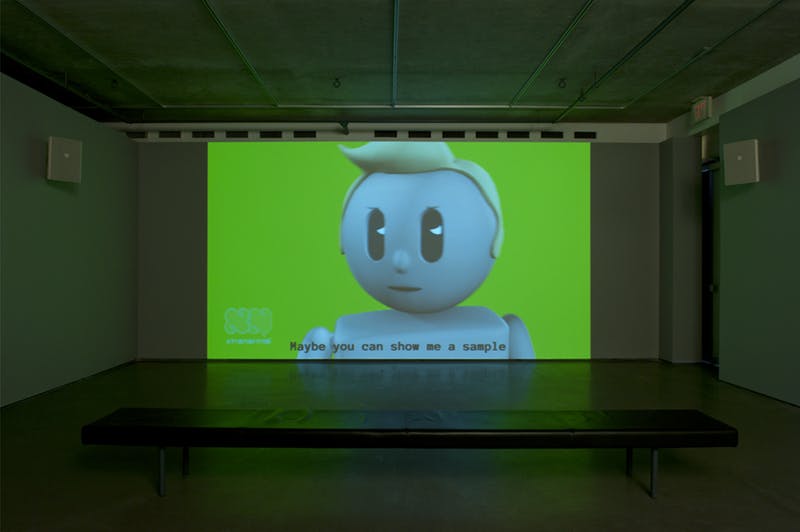 A single-channel video is projected on a wall in a darkened gallery space. The scene depicts a toy-like, animated man saying “Maybe you can show me a sample” against a bright green background.