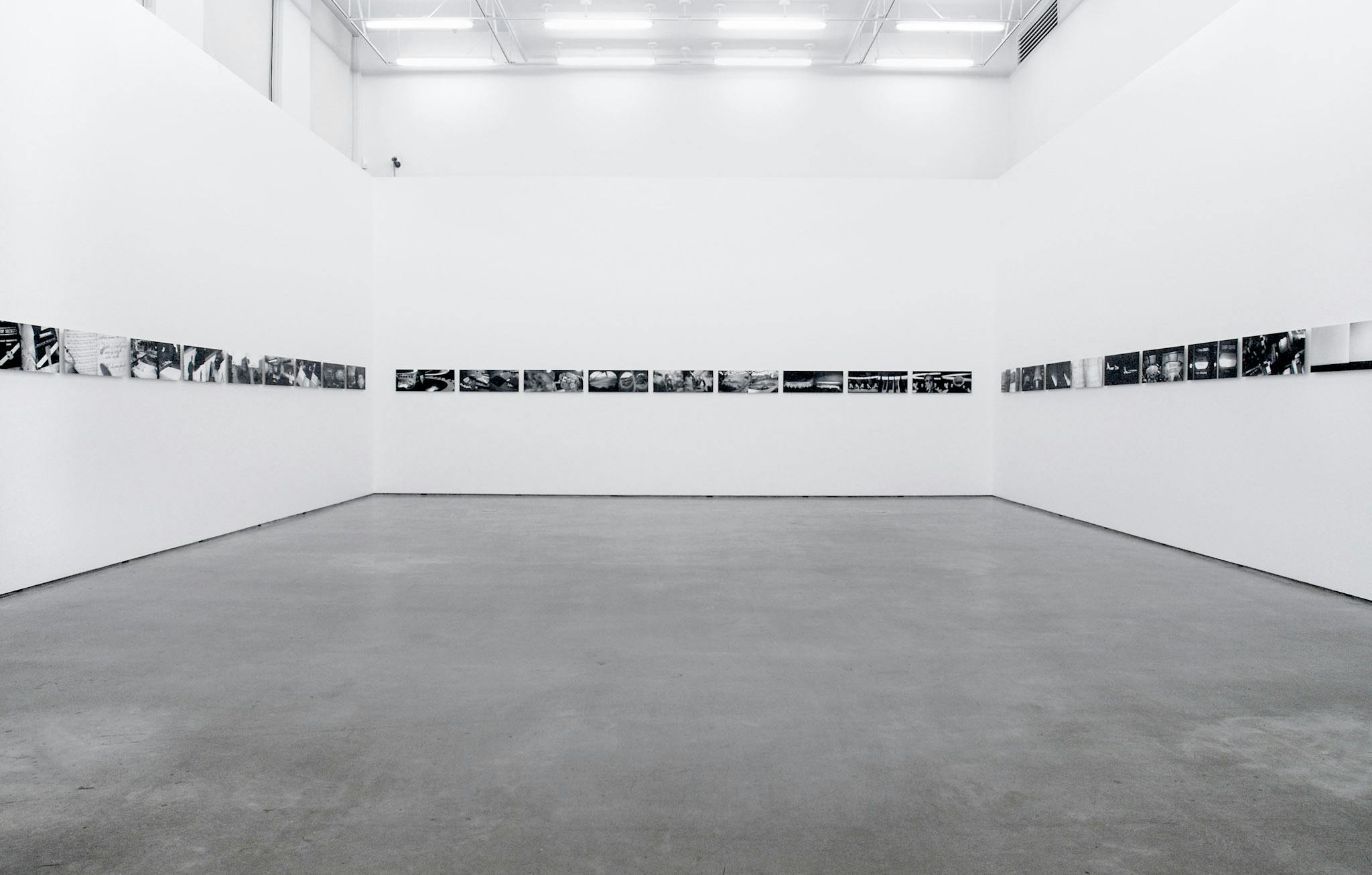 This is an installation shot of Kristan Horton’s art exhibition at CAG. Across three white walls, same-sized black and white photographs are mounted in a row. Many of those images depict landscapes. 