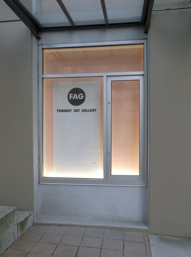 This photograph depicts one of the CAG window spaces. Behind a glass window on a pink wall, a large white filter is mounted. The flier says FAG, Feminist Art Gallery in black block fonts. 