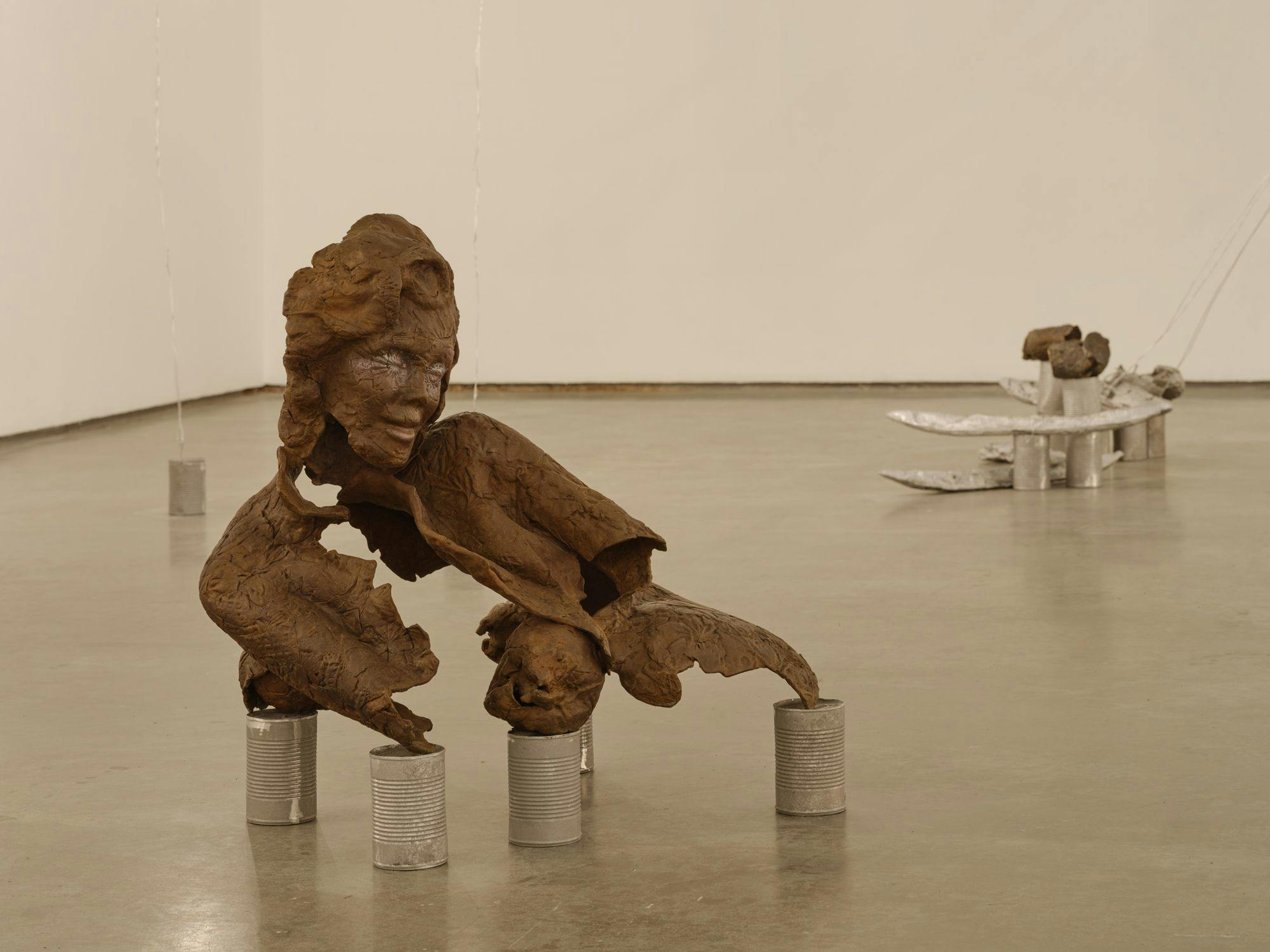 A bronze sculpture of a partial human figure sits on aluminum cans on a concrete floor. The sculpture's form appears to be precarious. Behind it are silver baguettes and aluminum cans.
