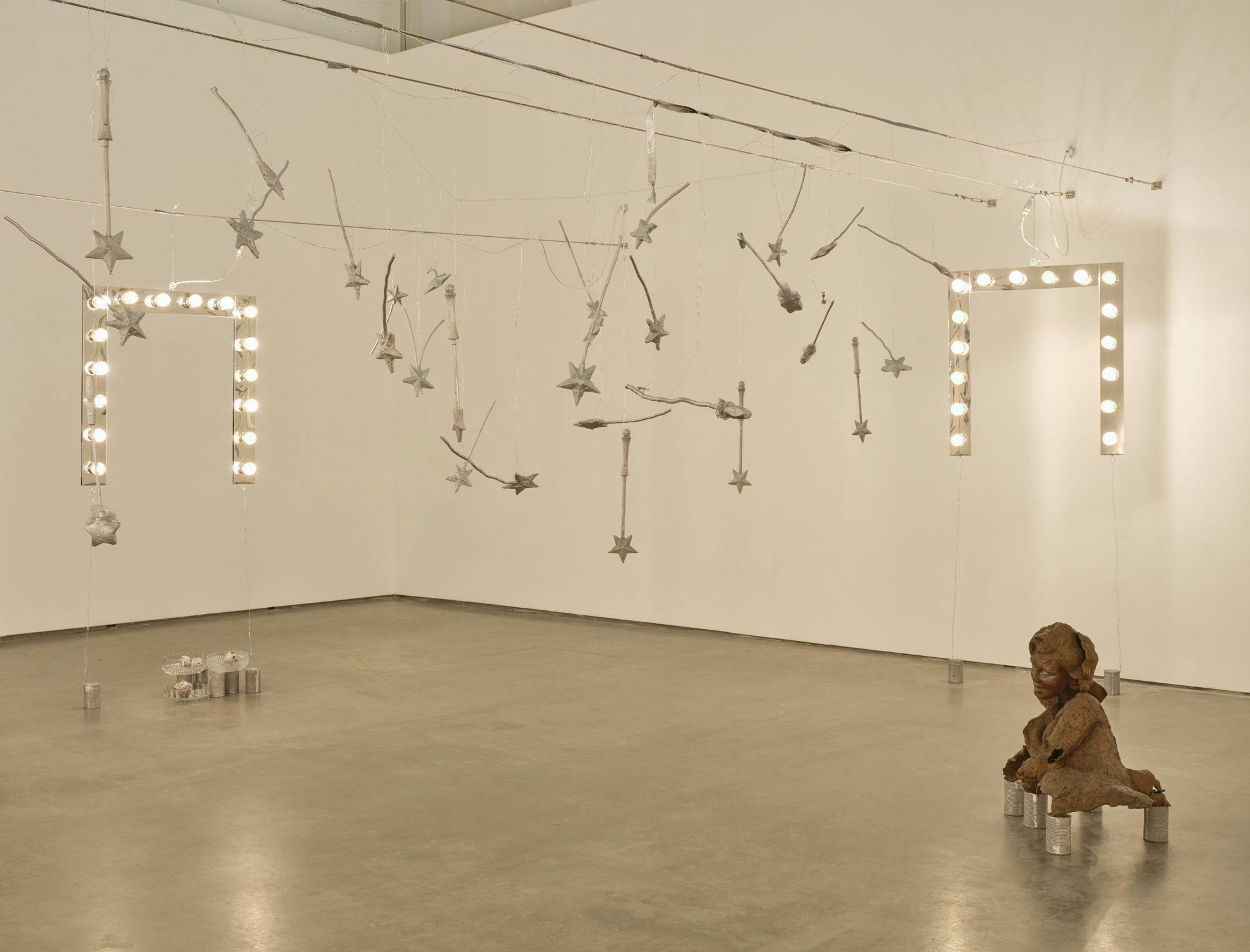 A wide view of vanity lights and silver magic wands hanging from wire. A bronze sulpture of a partial human figure sits on aluminum cans.