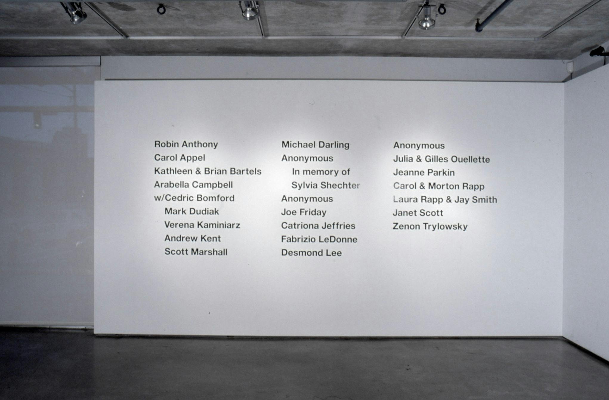 This is an installation shot of a gallery space. On a white wall, names of people are printed in a large black font. There are two listed as “Anonymous” in this list of names. 
