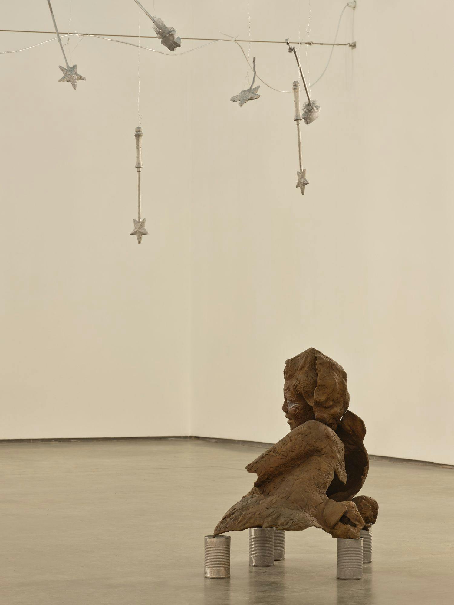 A bronze sculpture of a partial human figure sits on aluminum cans on a concrete floor. Above, silver magic wands hang from wire.