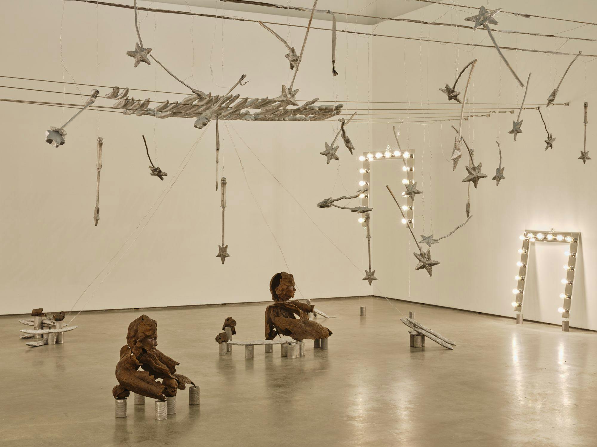 A wide view of several sculptures hanging from wire: magic wands, vanity lights and silver baguettes. Two bronze sulptures of partial human figures sit among aluminum cans and painted baguettes on the floor.