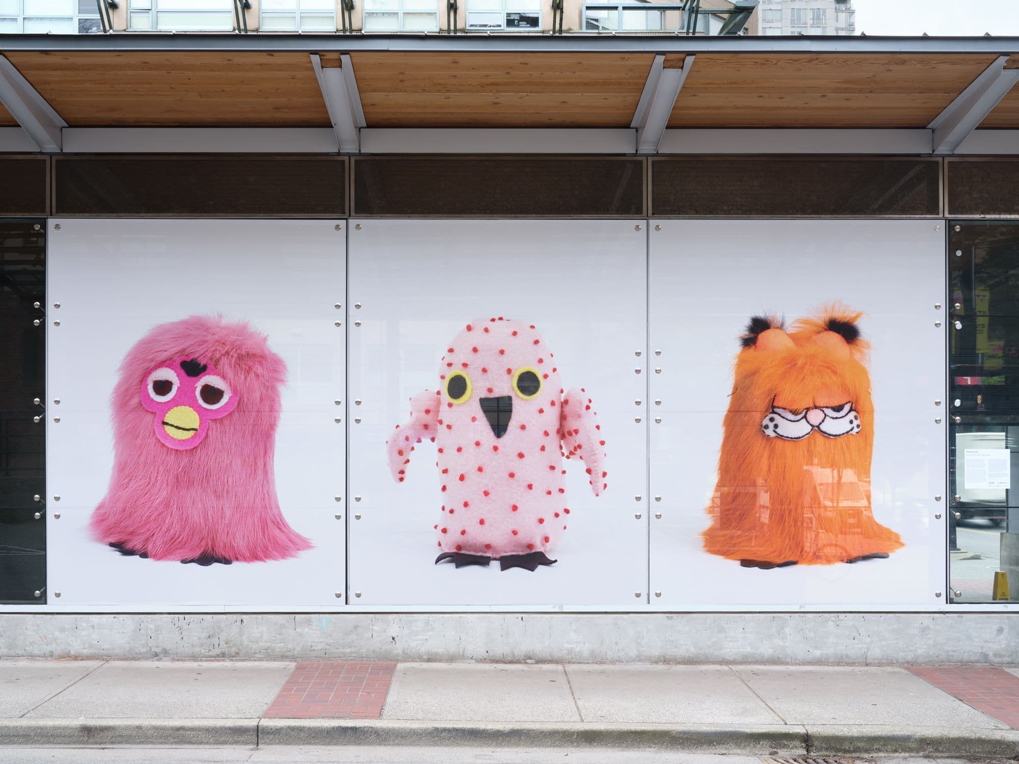 A frontal view of a SkyTrain station with photographs depicting Ookpik figures, or stuffed owl toys, in its windows. From left to right are a Furby-inspired Ookpik, one with no fur and a Garfield-inspired one.