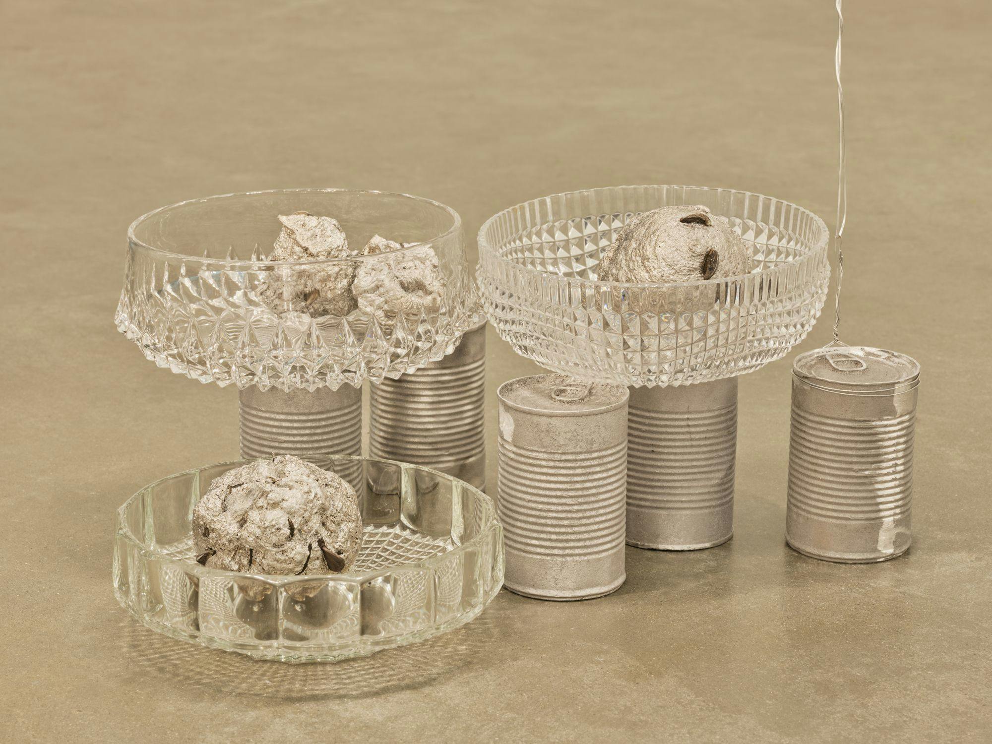 A sculpture comprised of three glass bowls and five aluminum cans. One of the cans is tied to wire that stretches out of frame.