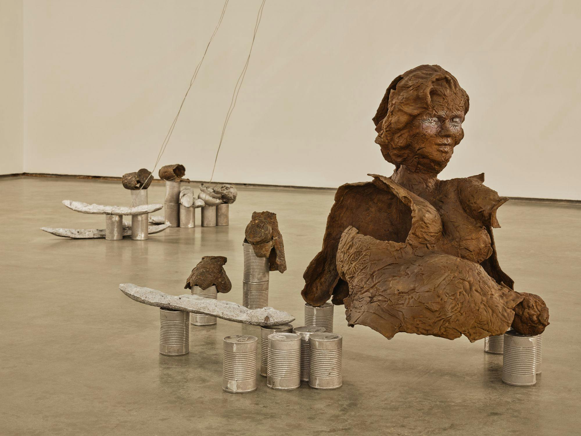 A bronze sculpture of a partial human figure sits on aluminum cans on a concrete floor. The sculpture's form appears to be precarious. Nearby are silver baguettes, aluminum cans and bronze cans that appear to be melting.