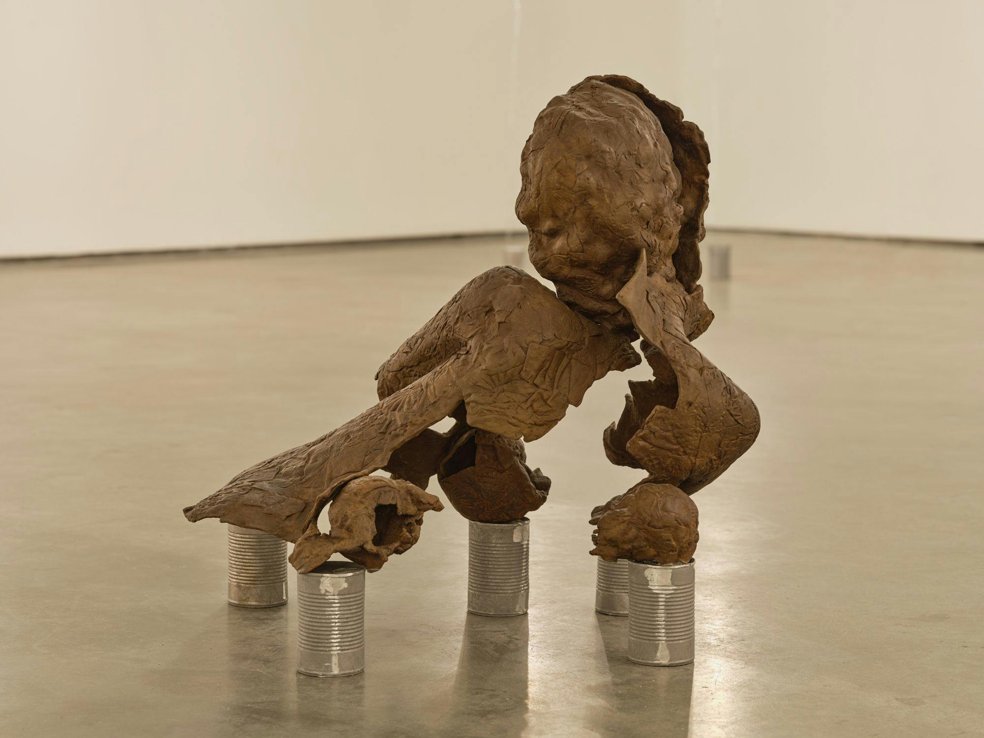 A bronze sculpture of a partial human figure sits on aluminum cans on a concrete floor. The sculpture appears to be disintegrating.