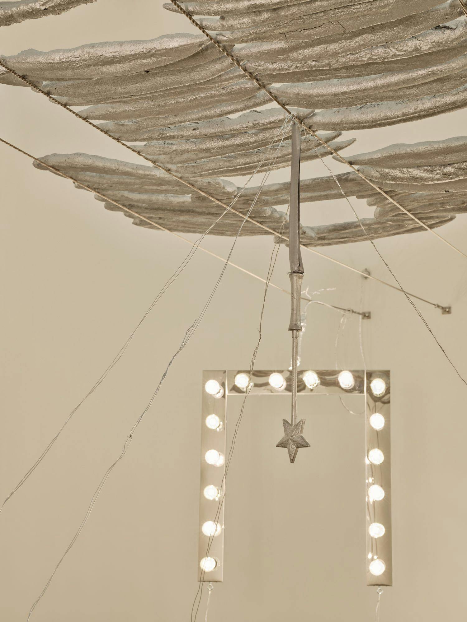 A silver magic wand, a vanity light and dozens of painted baguettes are held by wire fixed to the wall. Four wires hang from the silver wand and stretch out of frame.