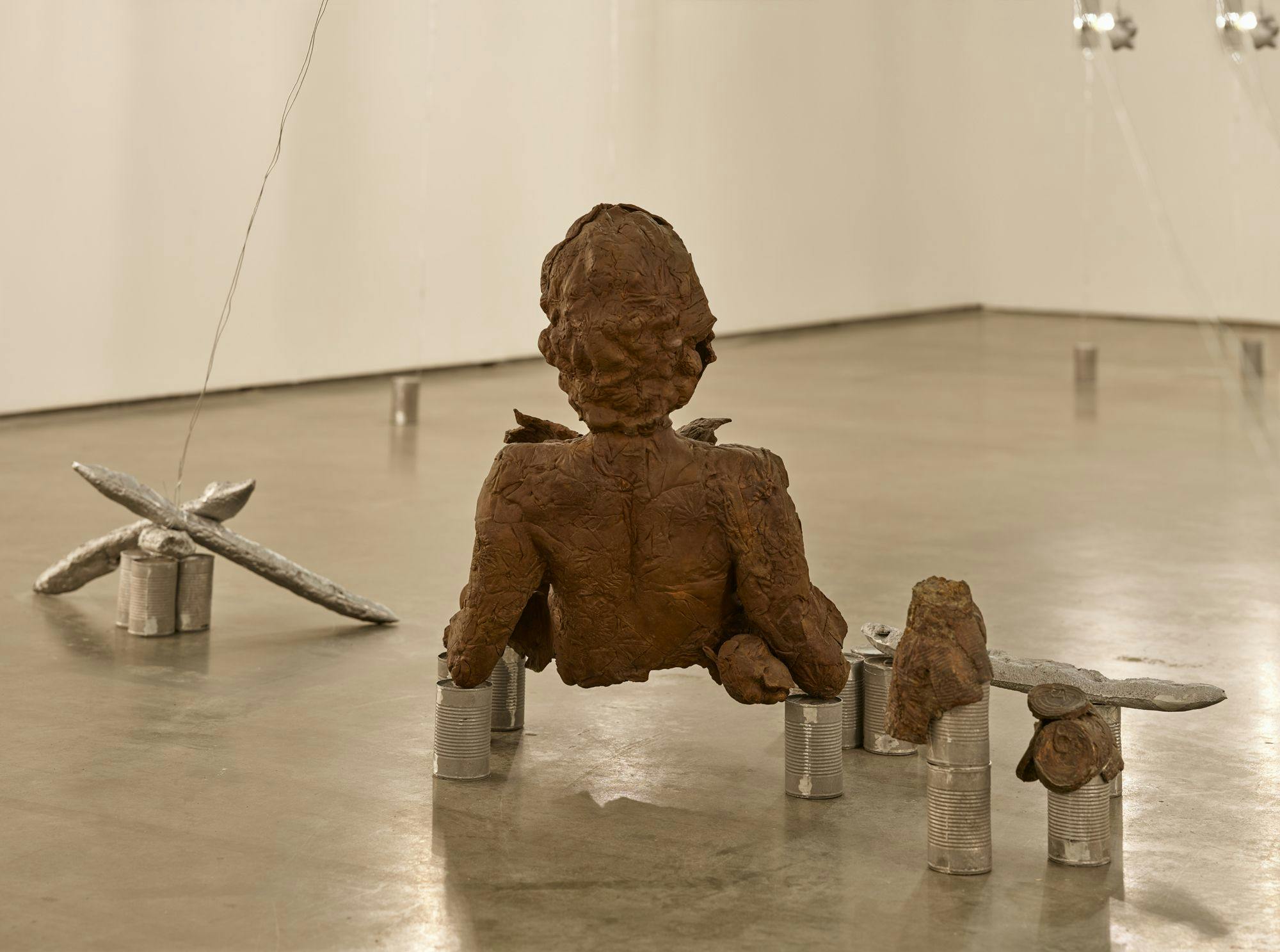 A bronze sculpture of a partial human figure sits on aluminum cans on a concrete floor. Nearby are more aluminum cans, silver baguettes, and bronze cans that appear to be melting.