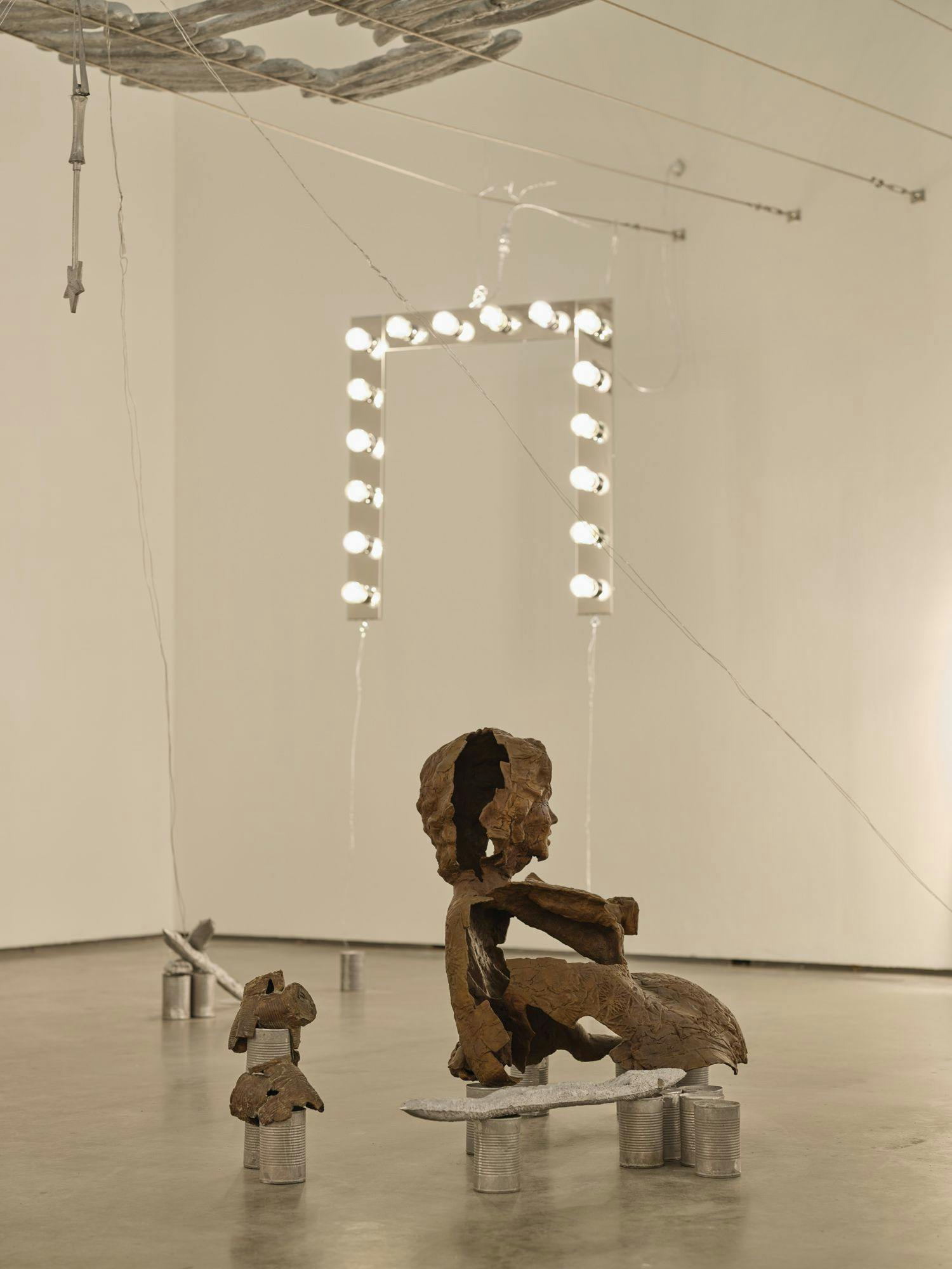 A bronze sculpture of a partial human figure sits among aluminum cans, baguettes and smaller bronze forms. Above, a magic wand, silver baguettes and a vanity light hang from wire.