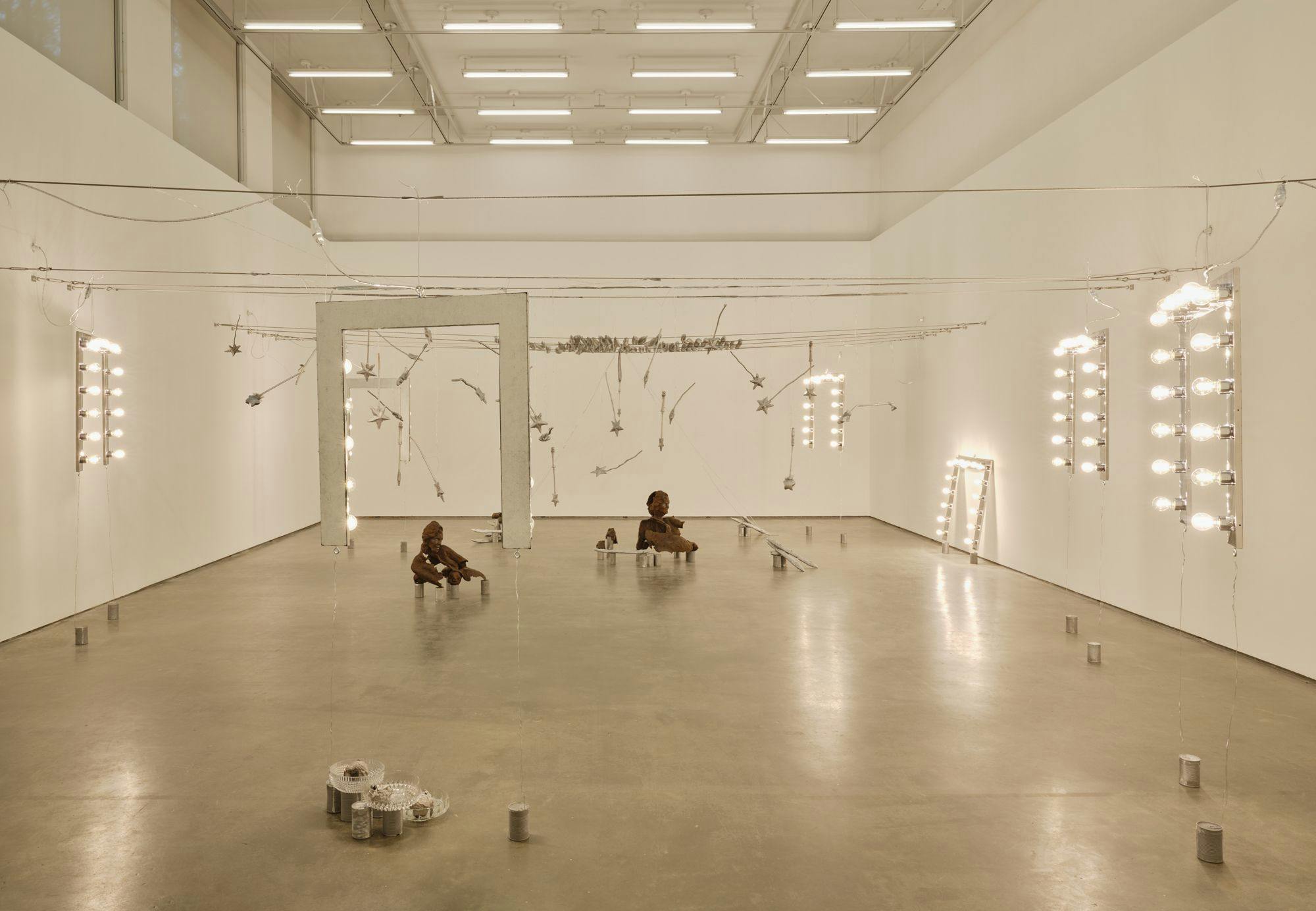 A wide view of several sculptures hanging from wire: magic wands, vanity lights and silver baguettes. Two bronze sulptures of partial human figures sit among aluminum cans, a vanity light, glass bowls, and painted baguettes on the floor.