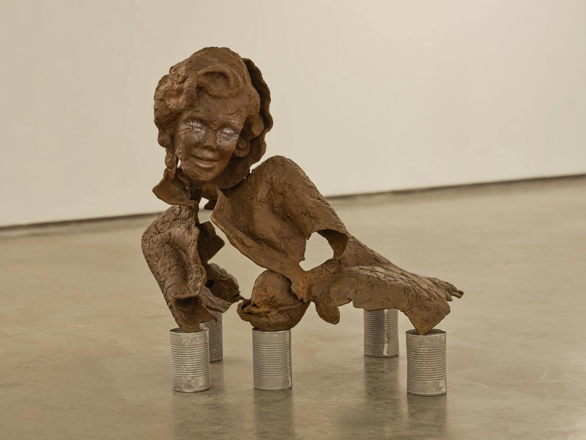A bronze sculpture of a partial human figure sits on aluminum cans on a concrete floor. The sculpture appears to be disintegrating.